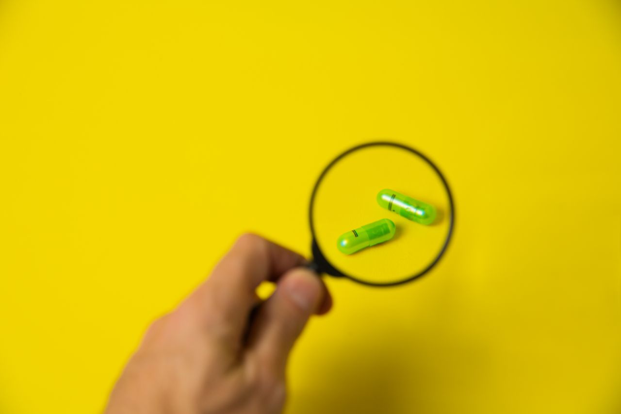 About From above of crop unrecognizable male holding magnifying glass over green pills on yellow background in studio